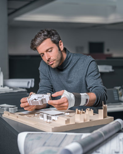 Architekt hält ein filigranes Modelhaus in den Händen und trägt eine Bort Bandage von Orthopädietechnik Nonnengäßer.